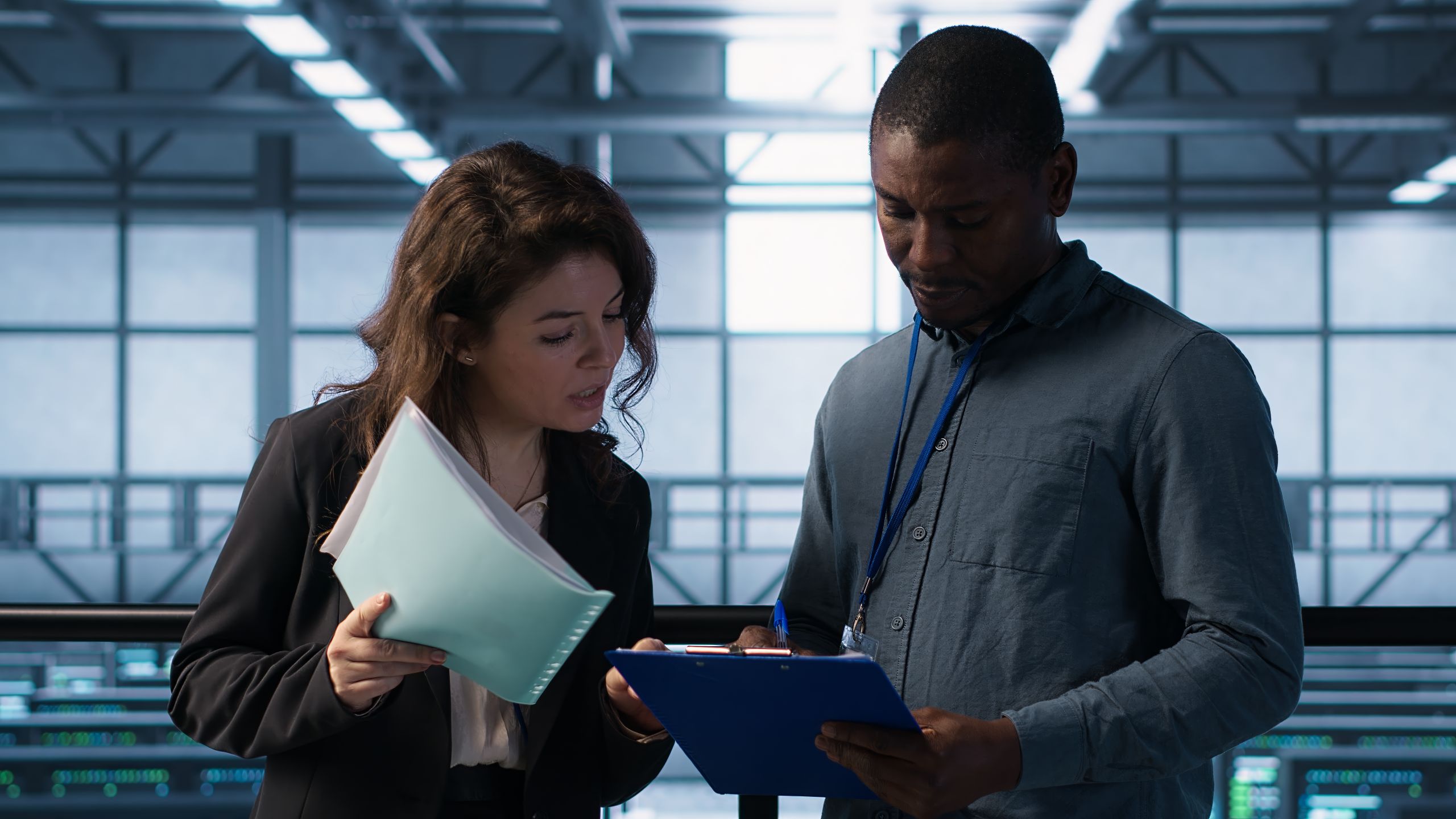 Two IT specialists holding clipboard and binder having discussion inside data center.