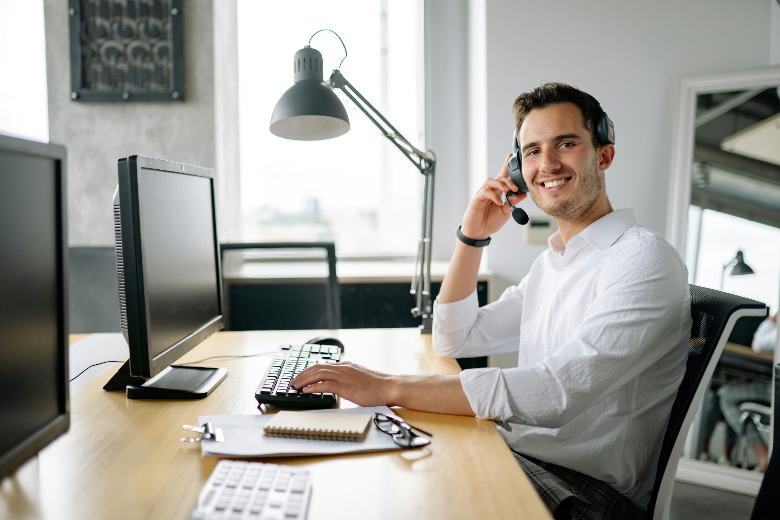 IT professional smiling in front of work station wearing headphones with a microphone.